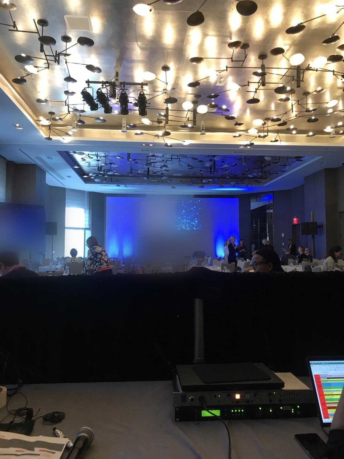 Corporate conference general session with branded stage screen and blue uplighting, viewed from the FOH tech position